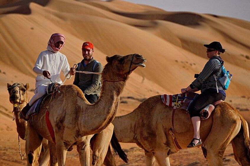 Tres personas montando camellos en un desierto con dunas de arena al fondo. 3