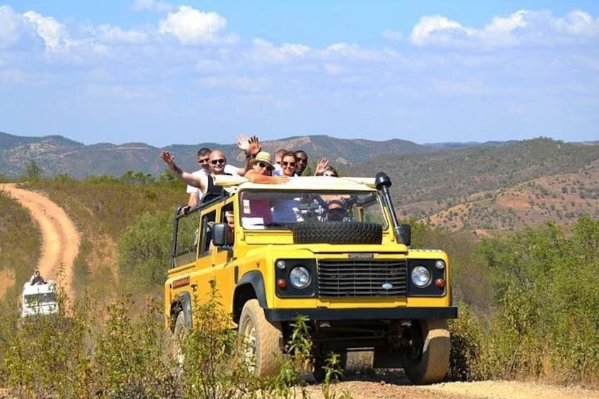 A yellow off-road vehicle with passengers on a dirt road surrounded by hills. 1