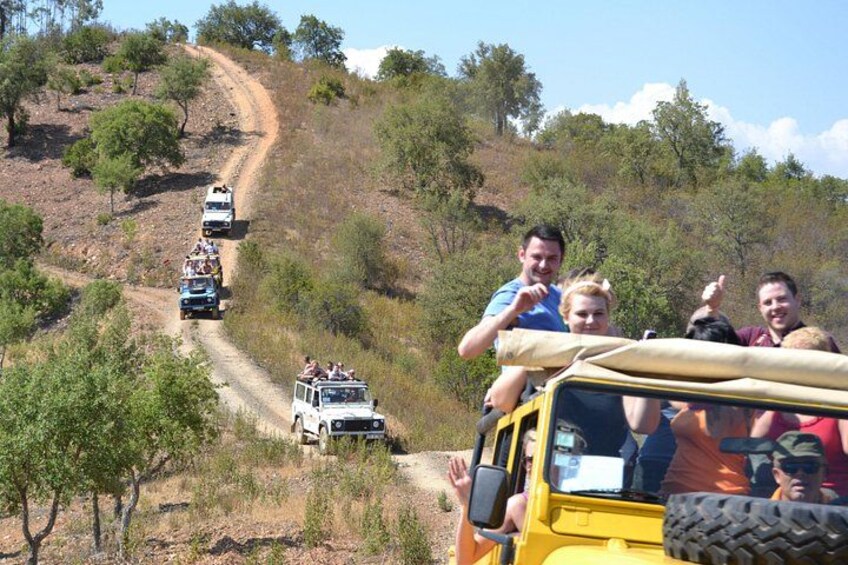 Een groep mensen op safari, voertuigen rijden over een stoffige weg door een droog, heuvelachtig landschap met schaarse begroeiing. 2