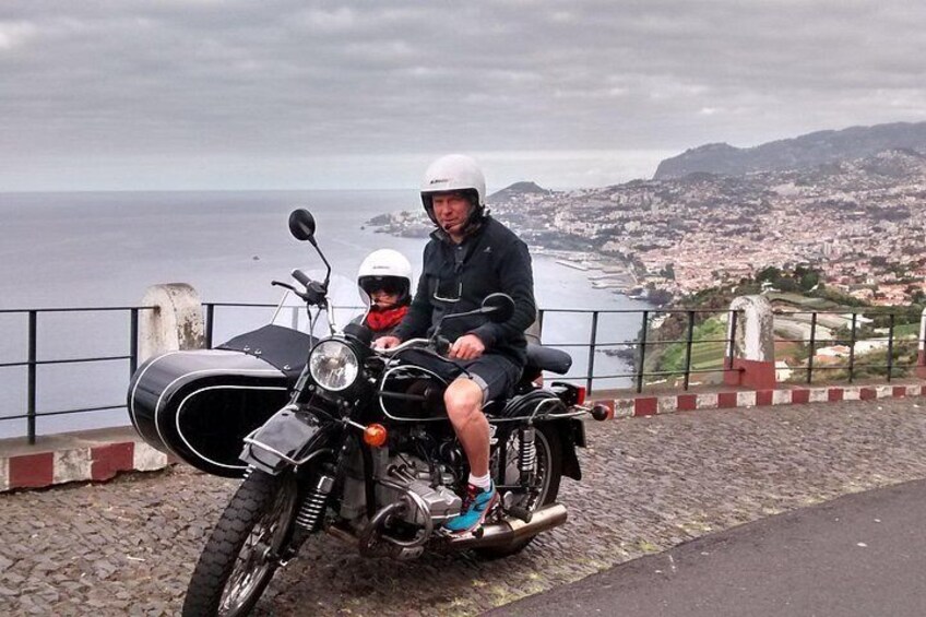 A motorcycle with a sidecar parked on a cobblestone path, overlooking a coastal cityscape with a body of water and hills in the background. 3