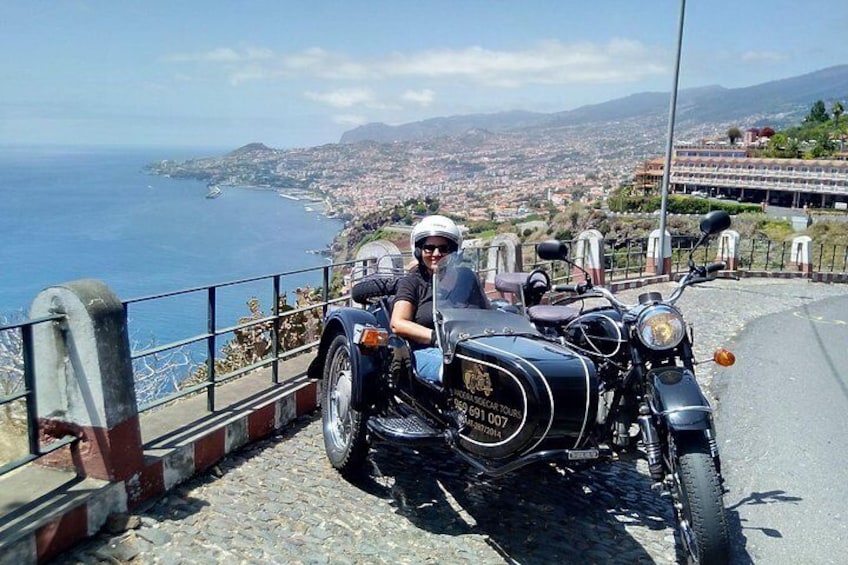 A person on a motorcycle with a sidecar parked on a road with a coastal view. 1