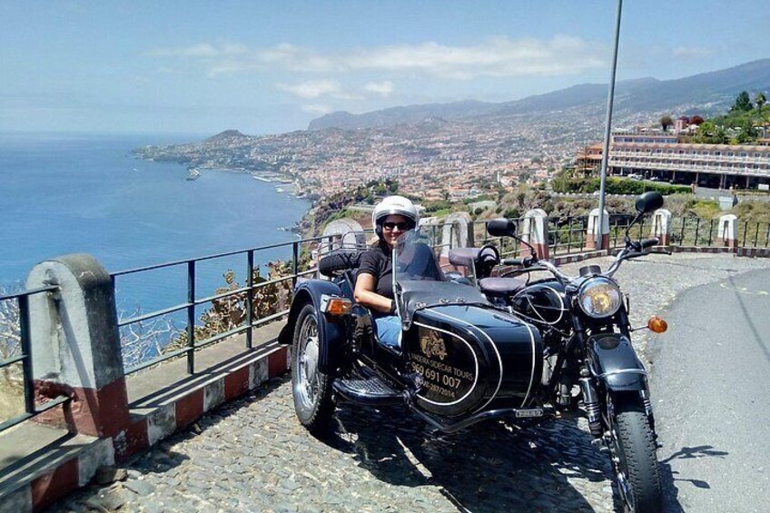 A person sitting on a motorcycle with a sidecar parked on a road with a coastal view. 1