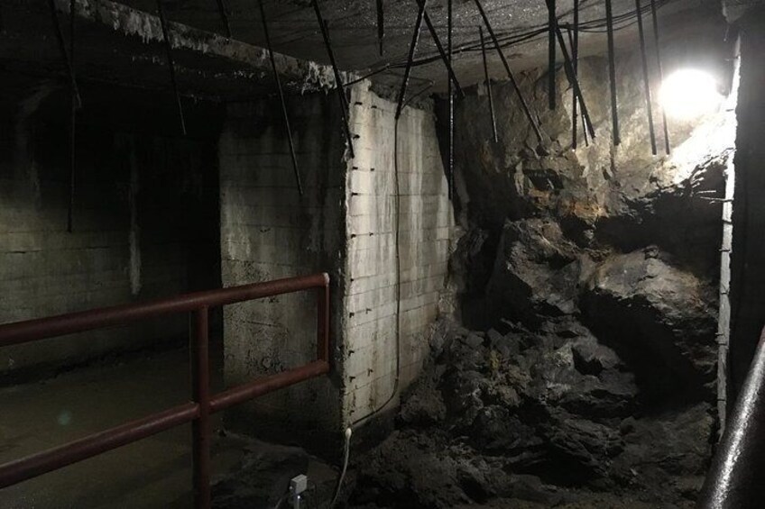An underground passage with concrete walls, a metal railing, and exposed rock formations. 1