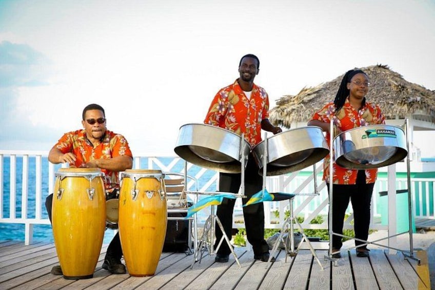Drie mensen bespelen drums op een houten terras met uitzicht op de oceaan. 2