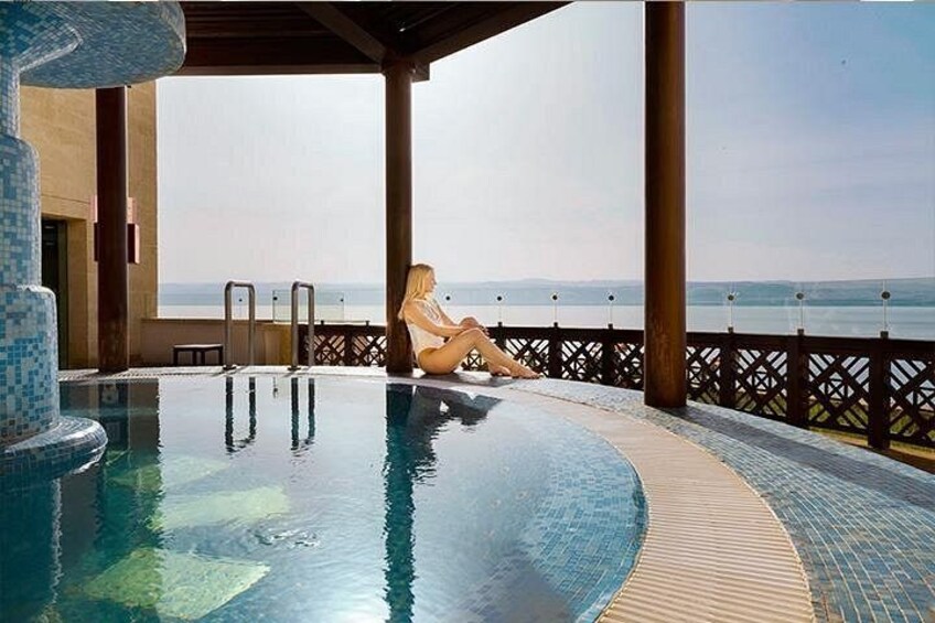 Una piscina con una persona sentada al borde, una terraza de madera y vista al mar. 2
