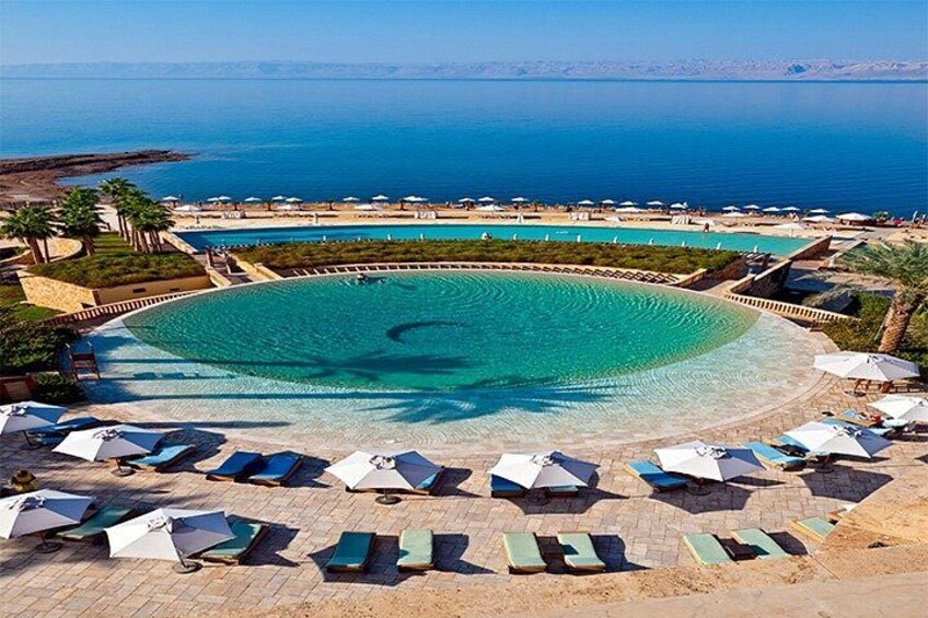 Una gran piscina circular con agua azul cristalina, rodeada de sillas de descanso y sombrillas, con vista a una playa y al mar. 2
