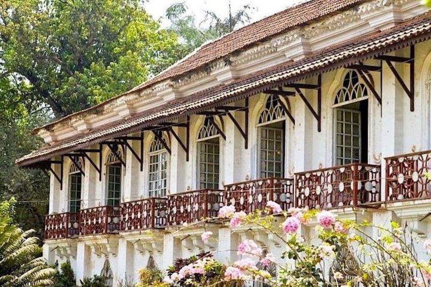 A colonial-style building with a balcony, surrounded by lush greenery and flowering plants. 2