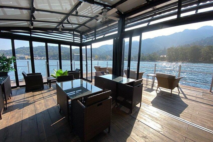 A covered outdoor deck with wooden flooring, glass tables, and chairs overlooking a lake with mountains in the background. 3