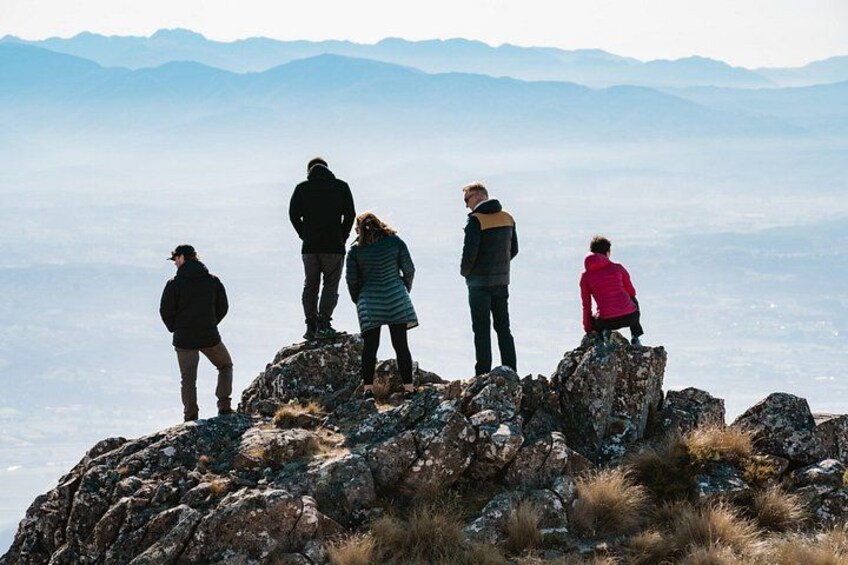 Een groep mensen staat op rotsachtig terrein, met een bergachtig landschap op de achtergrond. 2