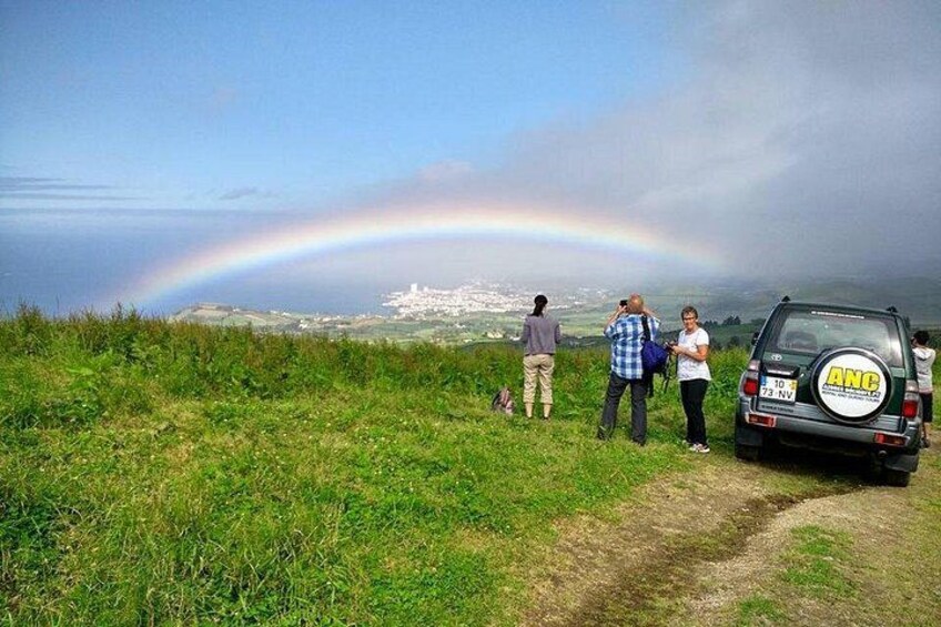 Un arc-en-ciel au-dessus d’un paysage côtier, avec des personnes qui l’observent, un véhicule garé et un ciel dégagé. 3