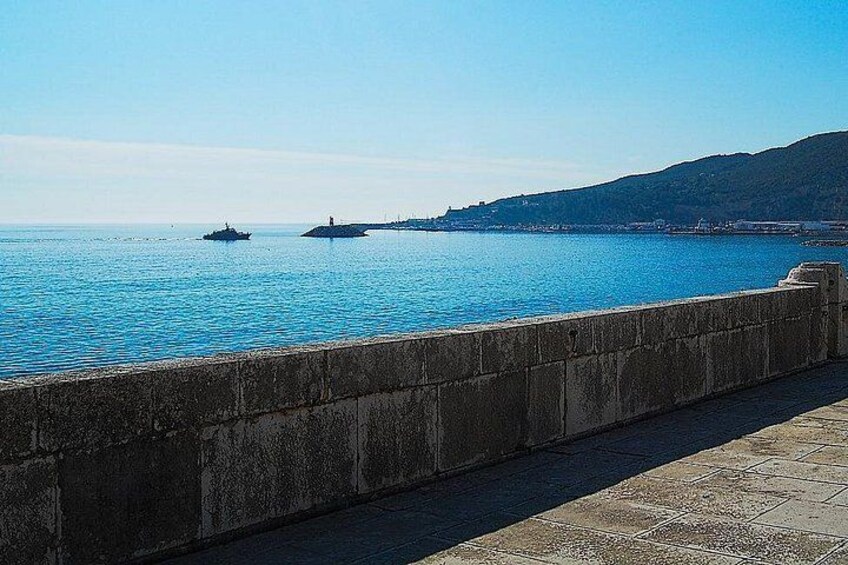 Paesaggio costiero con mare blu cristallino, un muro di pietra e un'isola all'orizzonte con un faro. 3