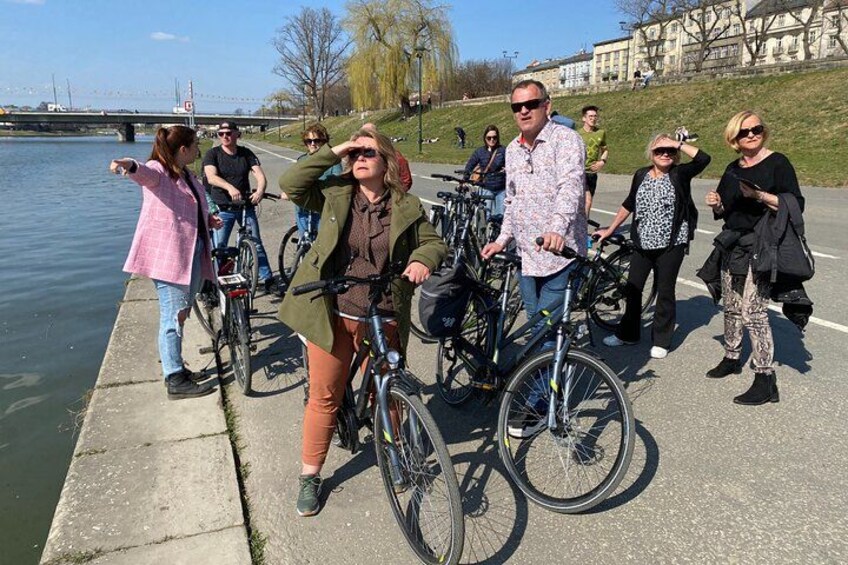Un groupe de personnes à vélo se trouve au bord d’une rivière, avec un pont en arrière-plan et des bâtiments de l’autre côté. 1