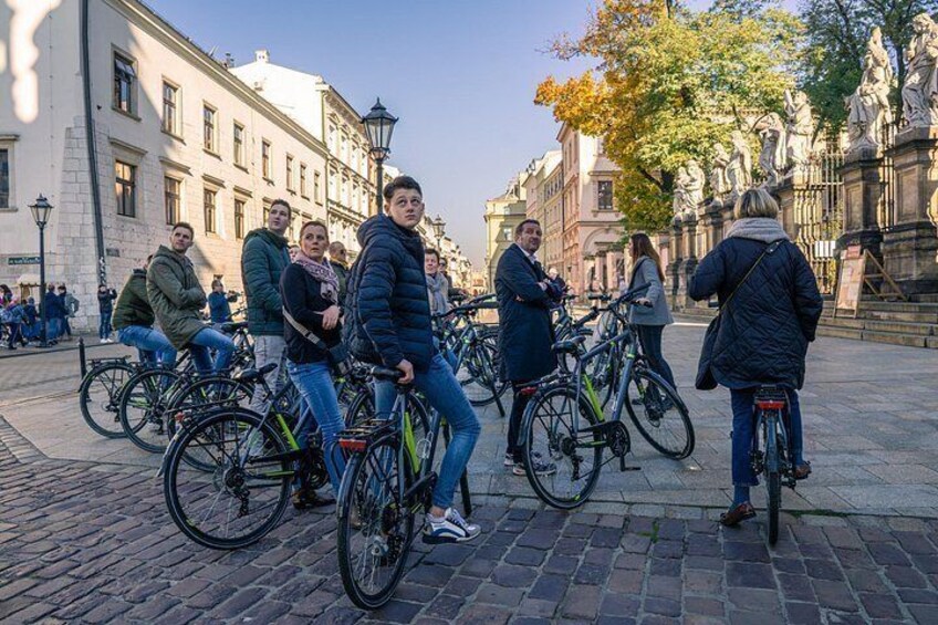 Een groep mensen op fietsen in een stedelijke omgeving met historische gebouwen en een standbeeld. 2