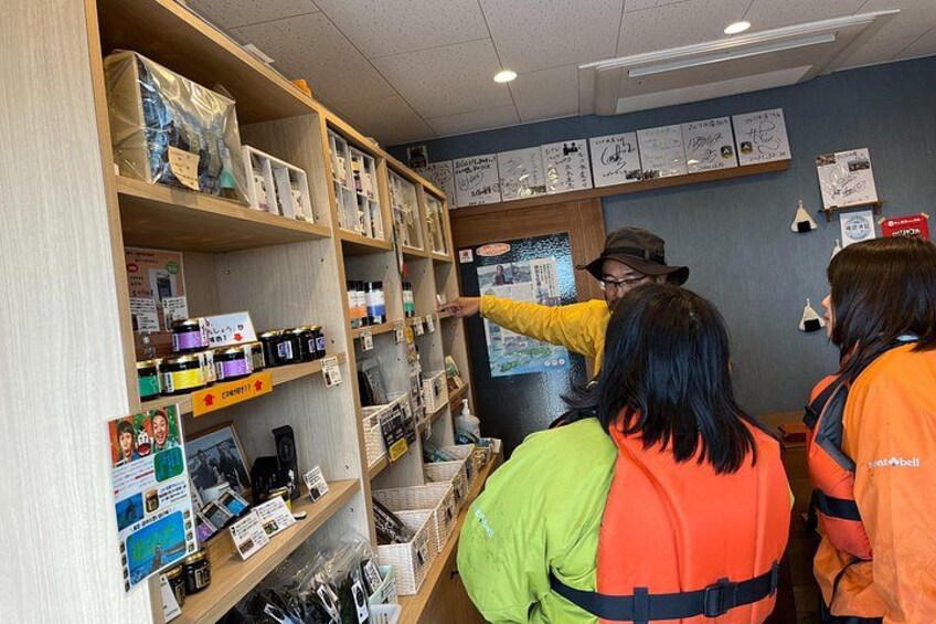 A person in a yellow jacket pointing at a shelf with various items, with two other people in orange jackets observing. 5