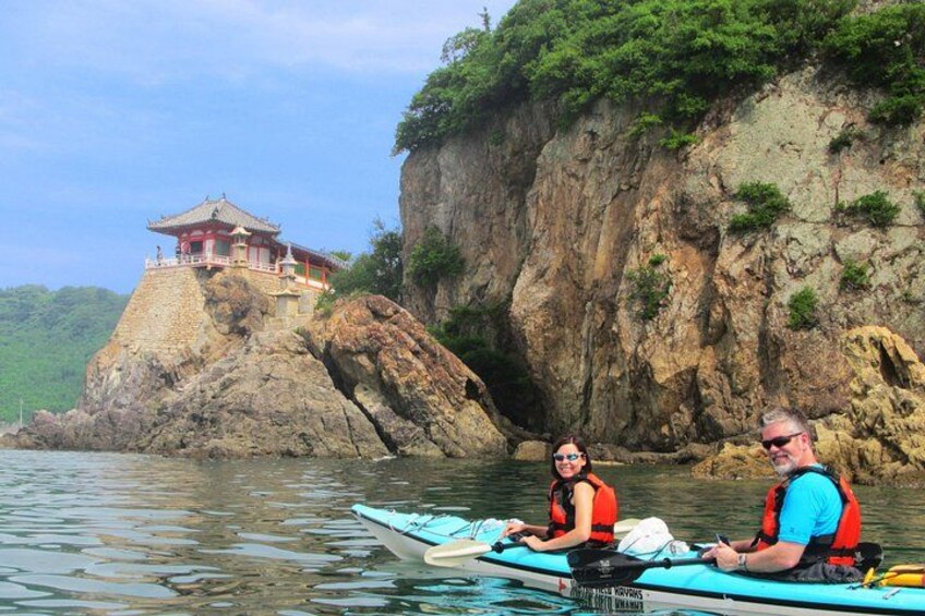 A couple kayaking on calm water with a traditional building on a rocky outcrop in the background. 1