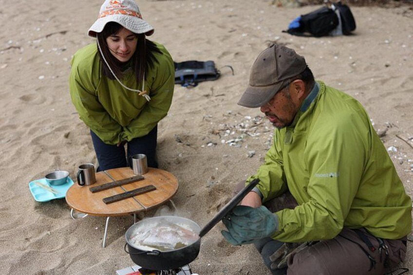 Two people cooking on a beach with a pot on a portable stove, a wooden board, and cooking utensils. 4