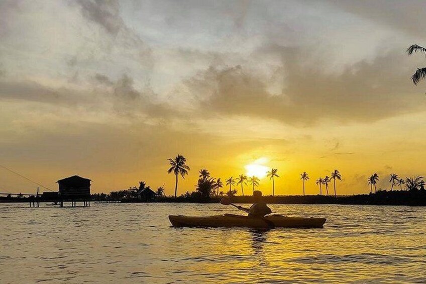 A sunset over water with a person paddling a canoe, palm trees, and a small building on the shore. 3