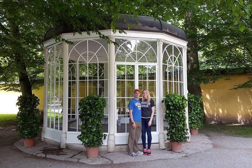 Ein gläserner Pavillon mit weißem Zierrat, umgeben von Topfpflanzen und Bäumen. 1