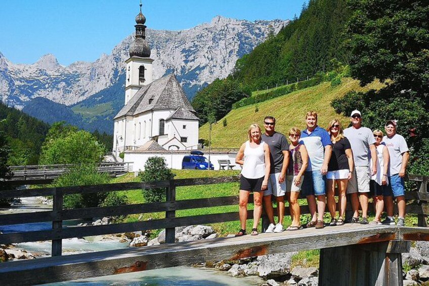 Eine Gruppe von Personen steht auf einer Holzbrücke, im Hintergrund eine Kirche und Berge. 4