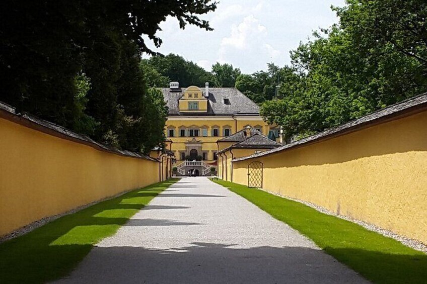 Ein Weg führt zu einem gelben Gebäude mit grauem Dach, umgeben von grünem Gras und Bäumen. 3