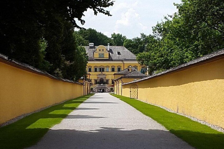 Ein Weg führt zu einem großen, gelben Gebäude mit grauem Dach, umgeben von grünem Gras und Bäumen. 3
