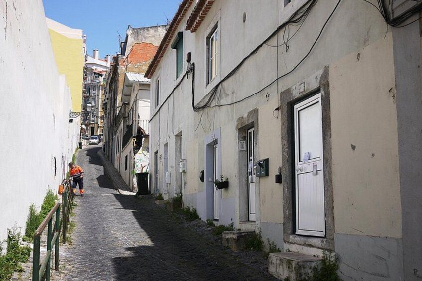 A narrow cobblestone street flanked by buildings with white and yellow facades, a person in orange attire, and a clear blue sky. 2