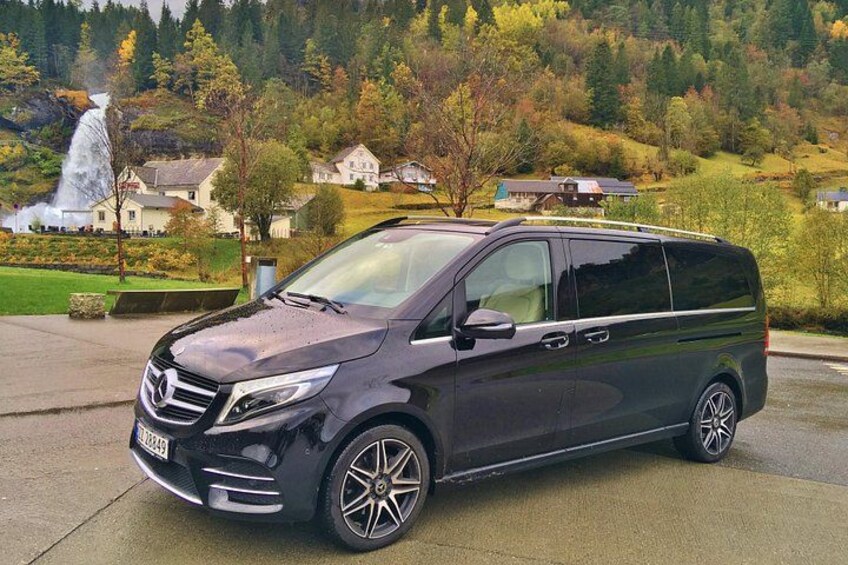 A dark-colored minivan parked on a concrete surface with a scenic background of a waterfall, trees, and houses. 2