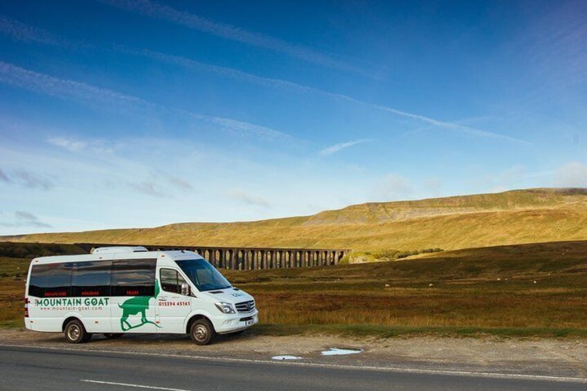 A white bus with 'Mountain Goat' branding parked on a road with a bridge in the background. 1