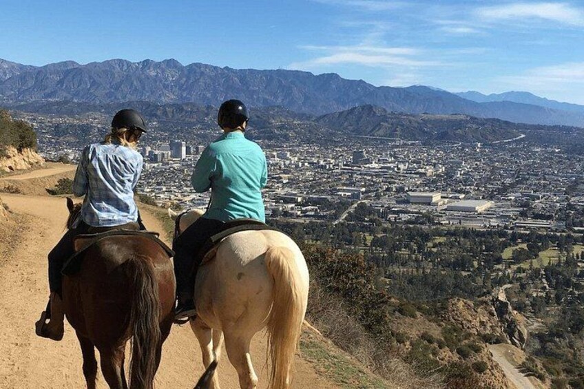 Dos personas montando a caballo por un sendero con vista a una ciudad y montañas. 1