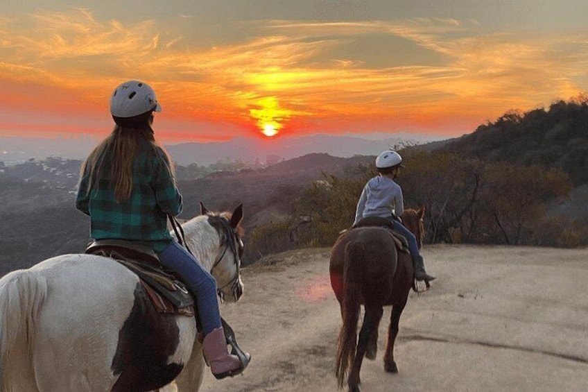Two people riding horses on a dirt path with a sunset in the background. 2