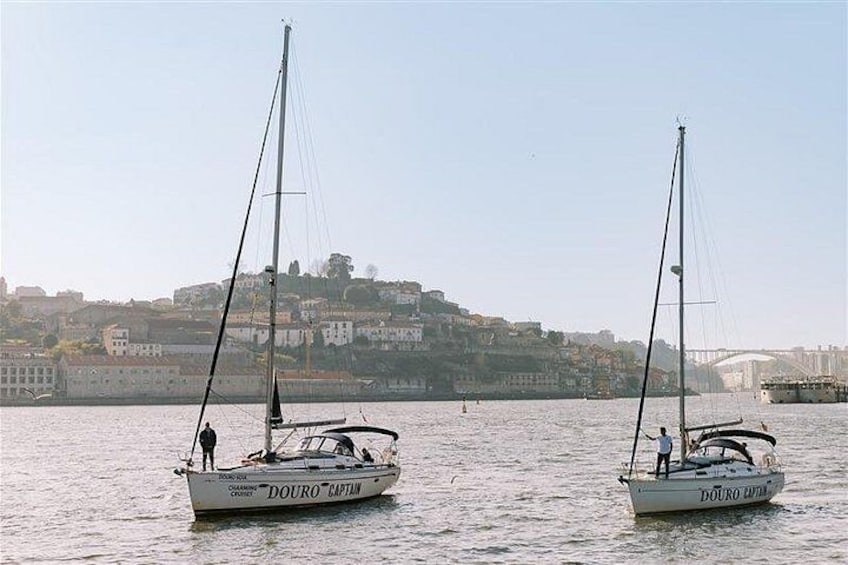 Two sailboats named 'Douro Captain' on calm water with a cityscape and bridge in the background. 3