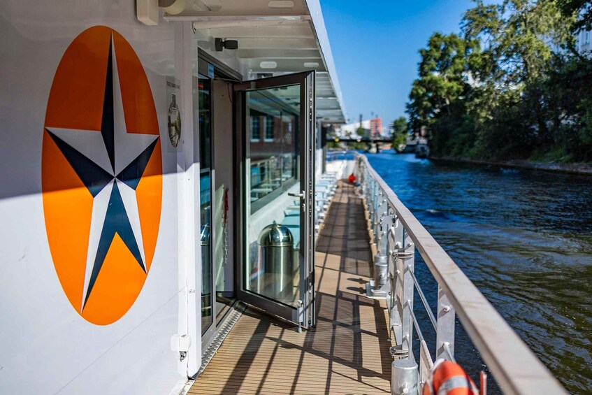 Un pont de bateau orné d’un logo en forme d’étoile, doté de portes vitrées et offrant une vue sur une rivière bordée d’immeubles et d’arbres. 4