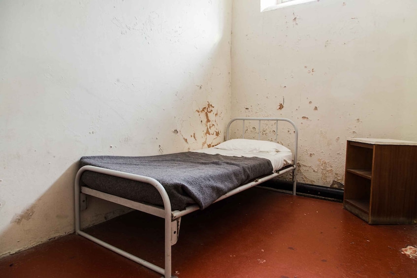 A small, single-bed room with peeling paint on the walls and a red floor. 2
