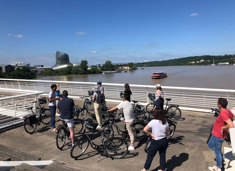 Des personnes se tiennent sur un pont, à côté de leurs vélos, et contemplent une rivière où naviguent des bateaux, avec un autre pont visible au loin. 1