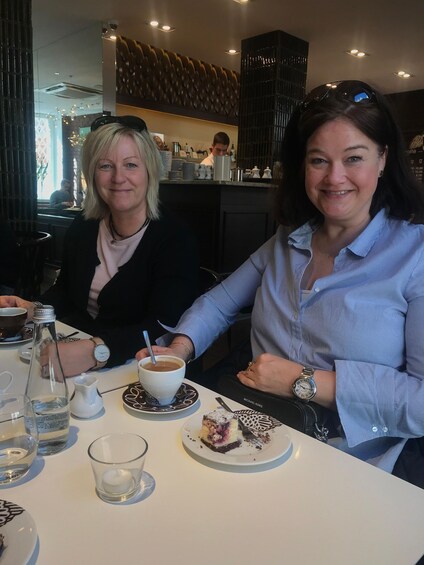 Two people sitting at a table with a coffee cup, a glass of water, and a slice of cake. 3