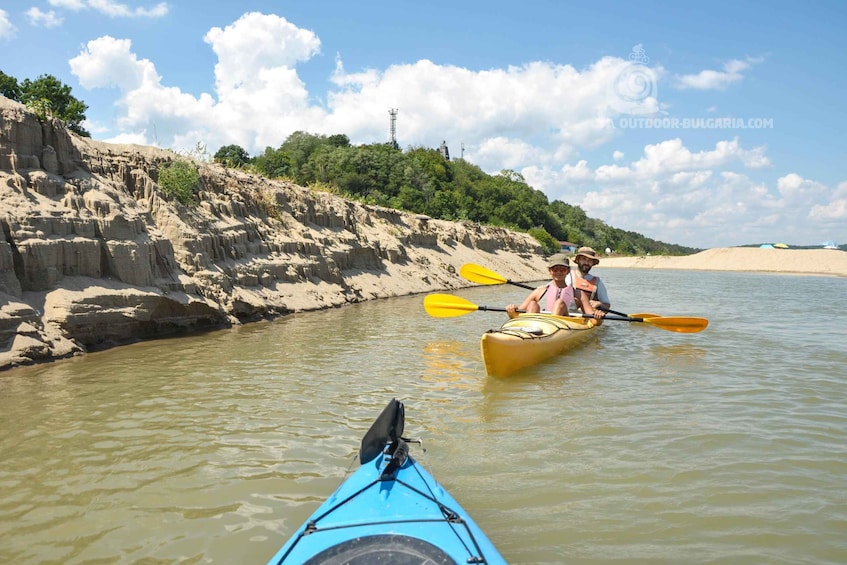 Zwei Personen paddeln mit ihren Kajaks auf einem Fluss, der von felsigen Ufern und einem Sandstrand im Hintergrund gesäumt wird. 1