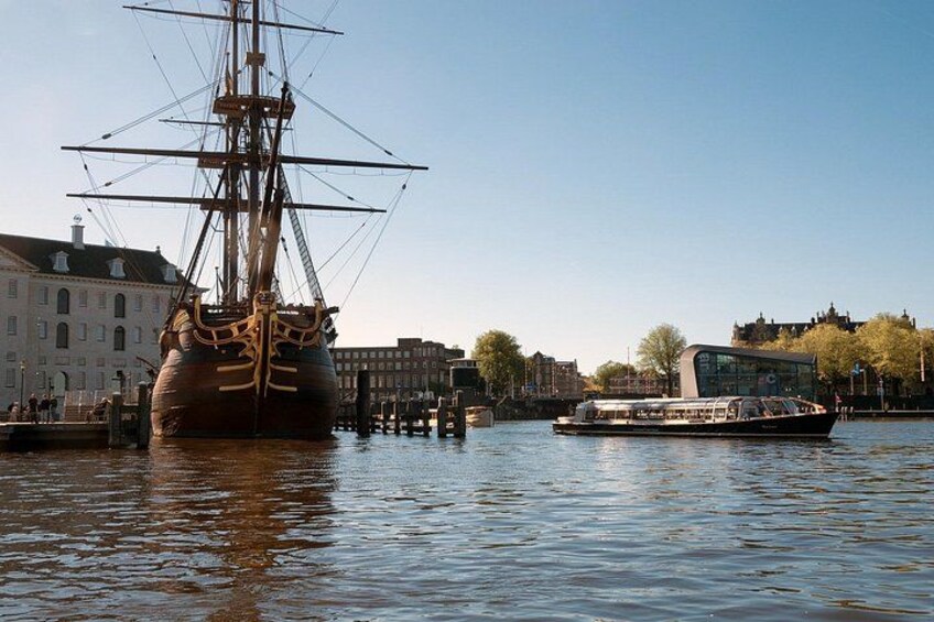 A large sailing ship docked at a harbor with modern buildings and a riverboat in the background. 4