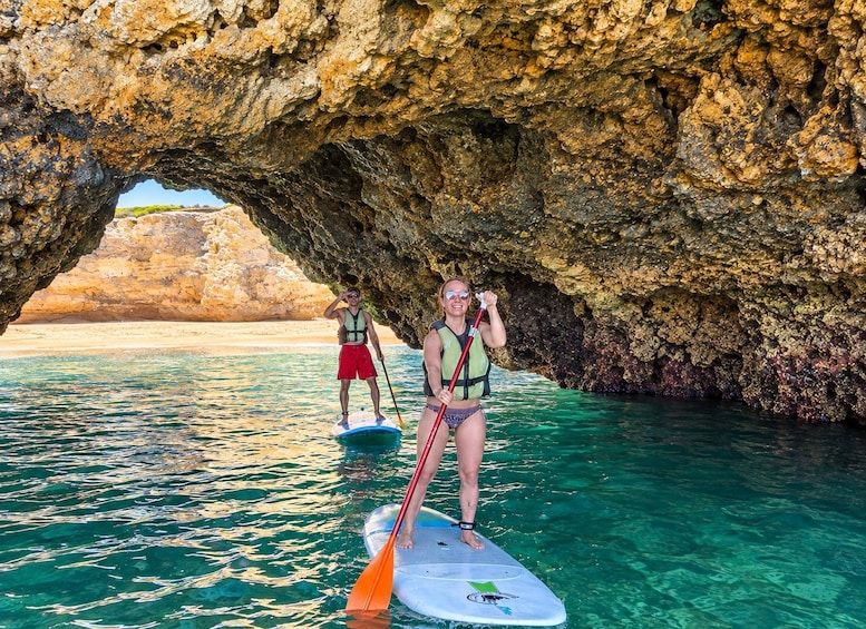 To personer padler på stand-up paddleboards nær indgangen til en klippefyldt hule, med krystalklart turkis vand. 1