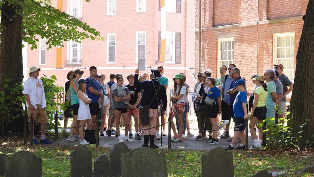 A group of people standing in a line near a cemetery with headstones, in front of a brick building with windows. 1