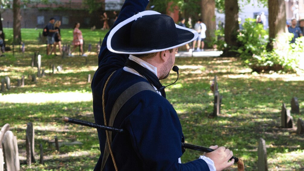 A person in historical costume holding a staff, standing in a park with trees and a fountain in the background. 2