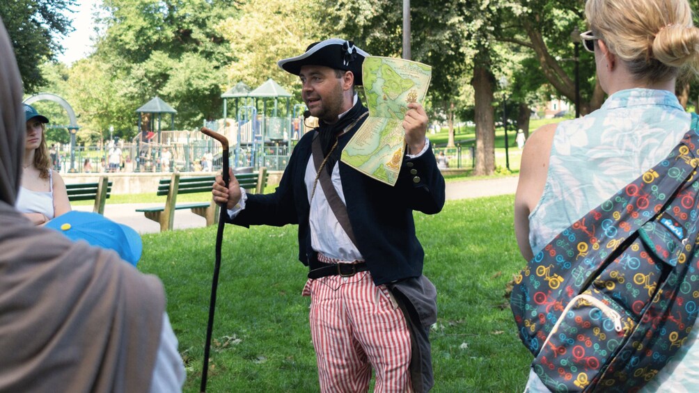 A person in historical costume holding a map, standing in a park with grass and trees. 4