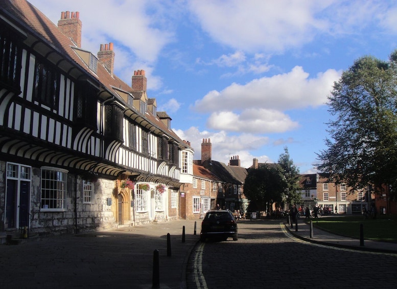 A cobblestone street lined with historic half-timbered buildings, a parked car, and pedestrians. 3