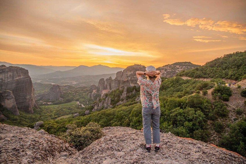 A person standing on a rocky outcrop overlooking a valley with mountains and greenery during sunset. 2