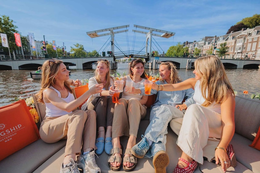 Een groep mensen zit op een boot en proost met drankjes, met een brug en gebouwen op de achtergrond. 3