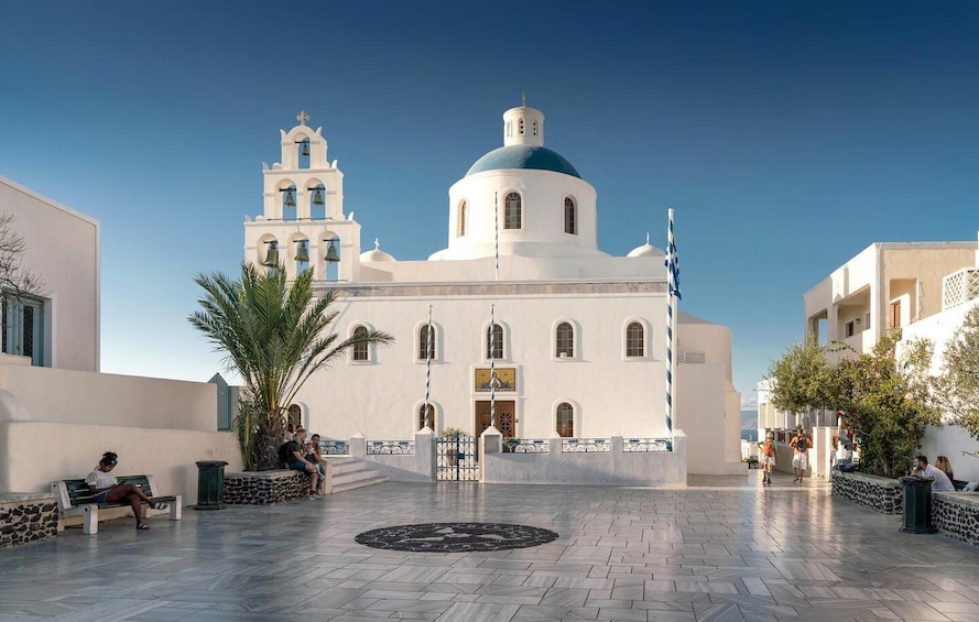Een witte kerk met een blauwe koepel en klokkentorens, een geplaveid plein met banken, en een helderblauwe lucht. 5