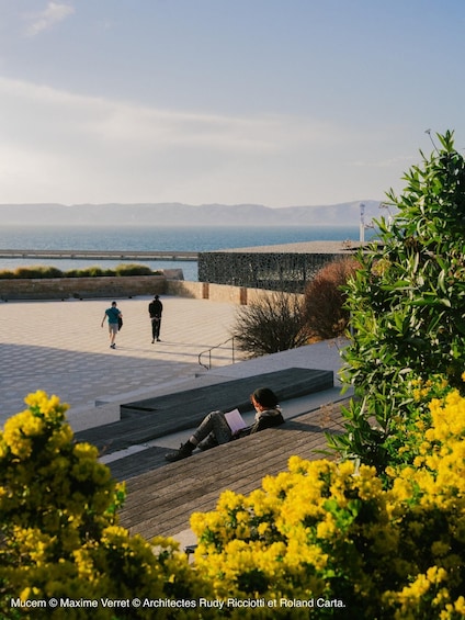 A coastal promenade with people walking and sitting, overlooking the sea and distant mountains. 5