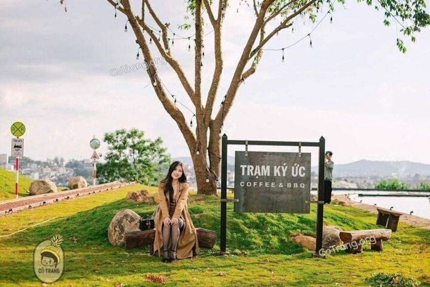 A person sitting under a tree with a sign for 'TRAM KY UÇ COFFEE&BQ' in a grassy area with rocks and a body of water in the background. 2