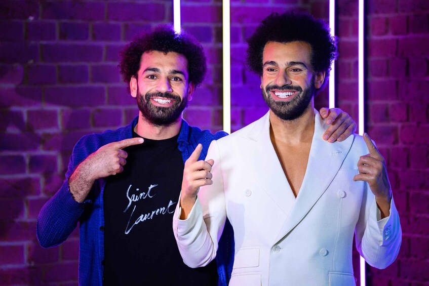 Two men standing in front of a brick wall with neon lights. One man wears a black shirt with white text, and the other wears a white suit. 5
