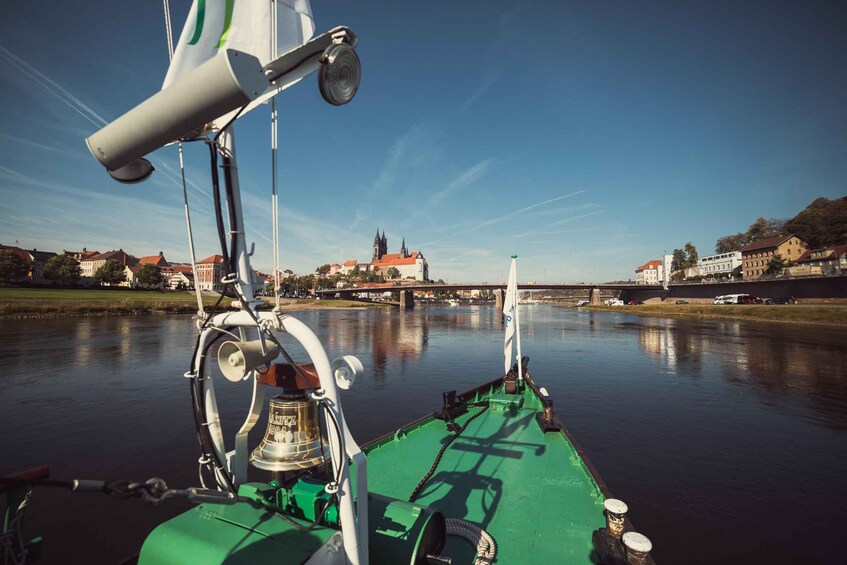 A green boat with a bell and a flag on the bow, a calm river, and a bridge with buildings in the background. 2