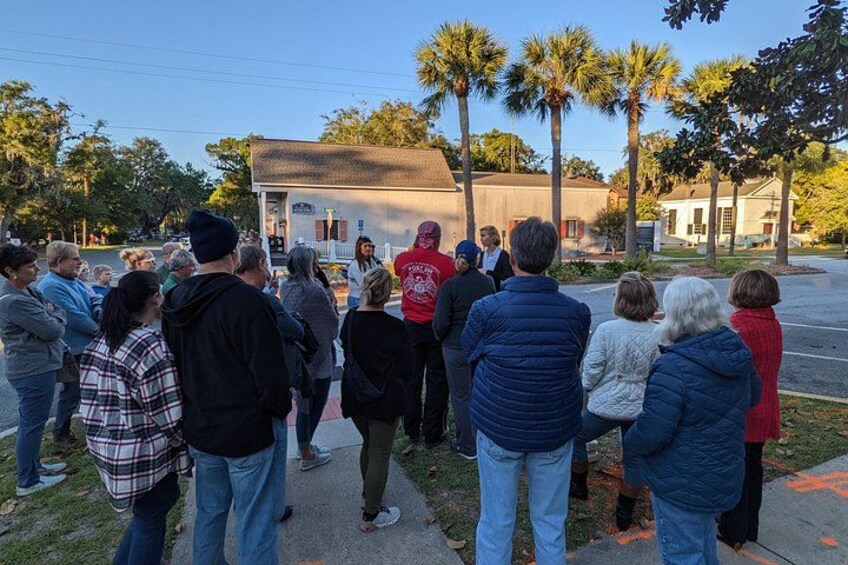 Un grupo de personas reunidas al aire libre, frente a un edificio con techo rojo, palmeras y una calle al fondo. 2